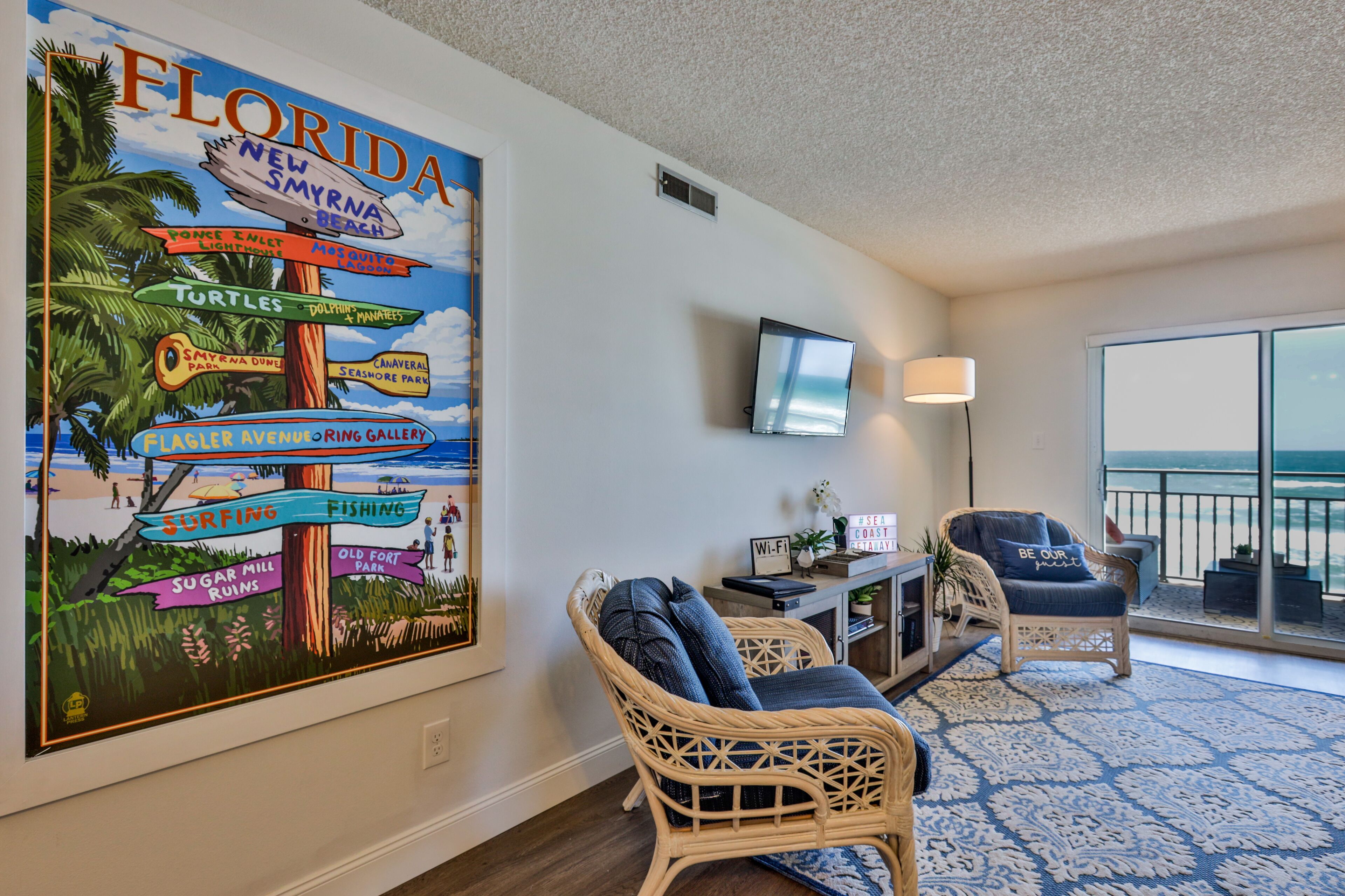 Ein Zimmer mit einem großen Schild im Florida-Stil, einem Fernseher, Korbstühlen, einer Lampe und Blick auf den Ozean.