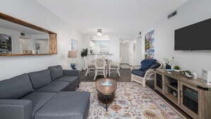A modern living room with a grey sectional sofa, a round coffee table, and a patterned rug. There is a dining area with wicker chairs and a white table. A television is mounted on the wall, and a bookshelf is visible.
