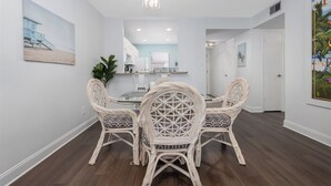 A dining area with wicker chairs and a glass table, white walls, and a beach painting.