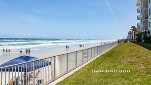 A beach with a fence, a grassy area, and a building with balconies.