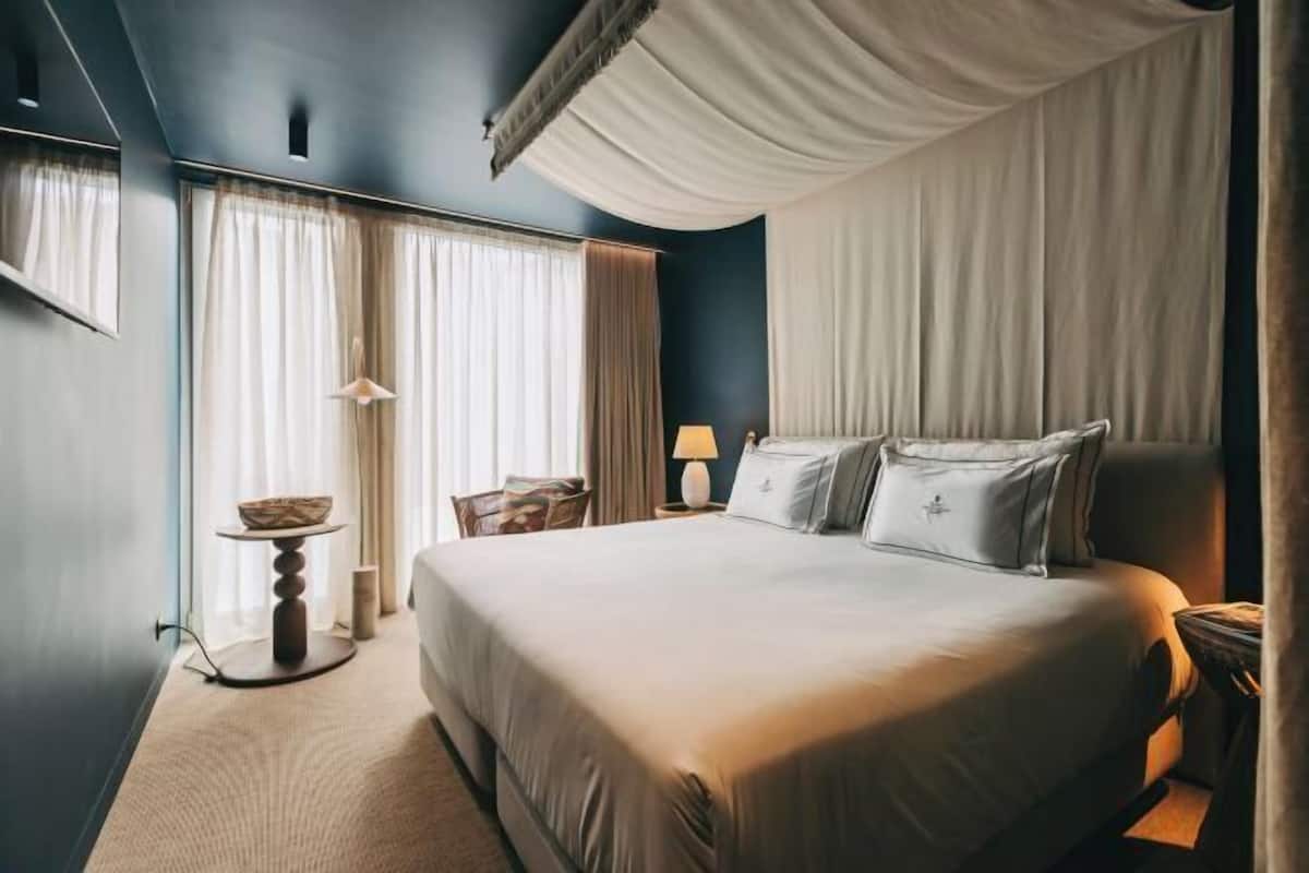 A hotel room with a large bed, a bedside table, a chair, and a window with curtains.