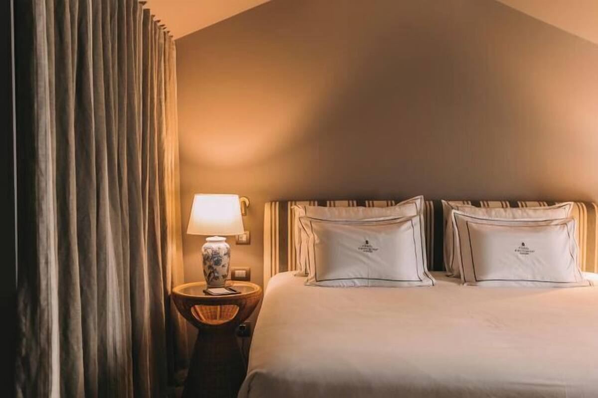 A hotel room with a bed, pillows, a nightstand with a lamp, and curtains.