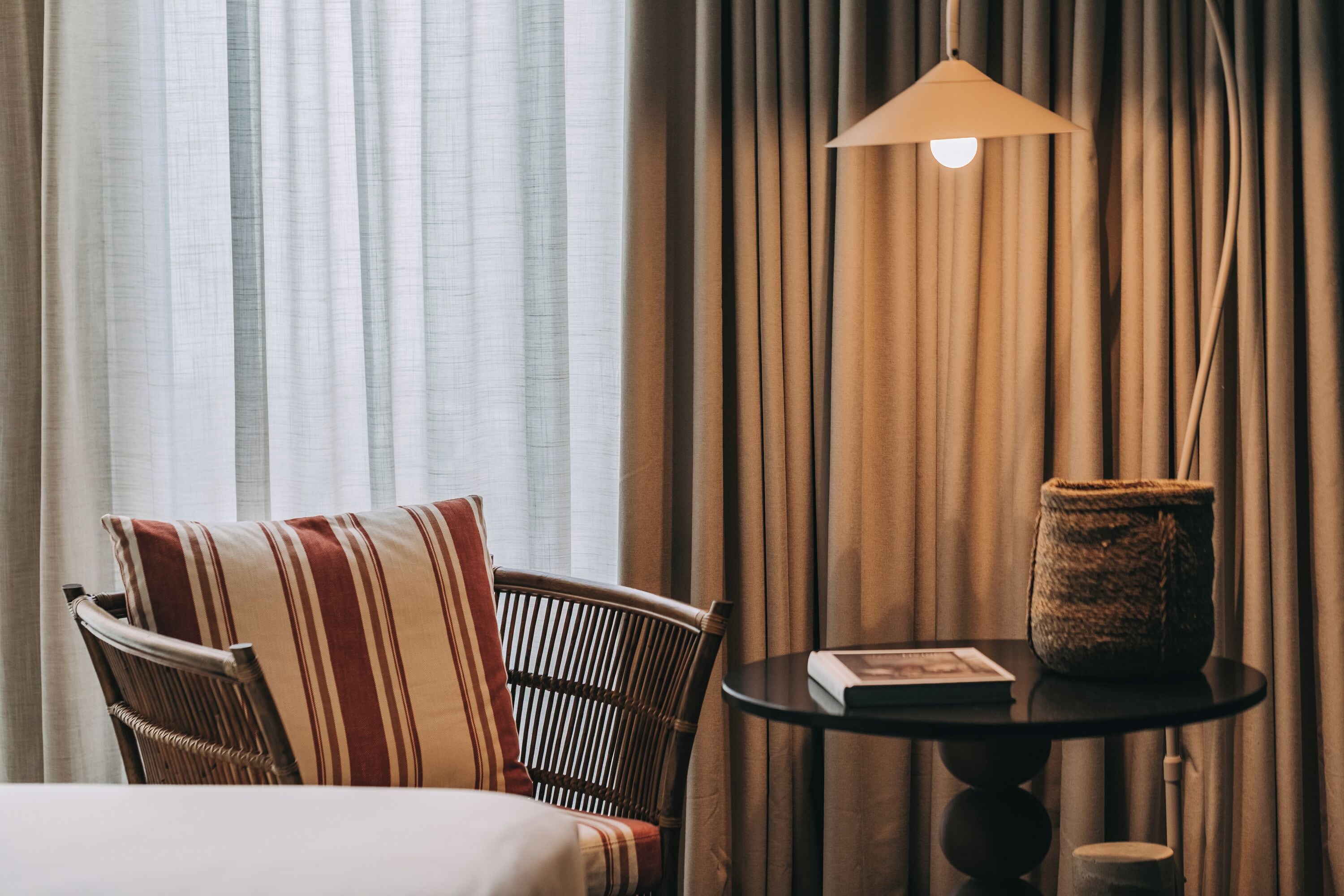 Quarto de hotel com uma cama, uma cadeira de vime com almofadas listradas, uma mesa redonda com um livro e uma cesta de palha, e um abajur.