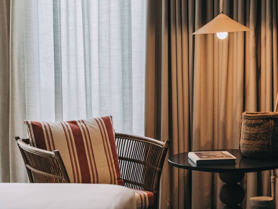 Quarto de hotel com uma cama, uma cadeira de vime com almofadas listradas, uma mesa redonda com um livro e uma cesta de palha, e um abajur.