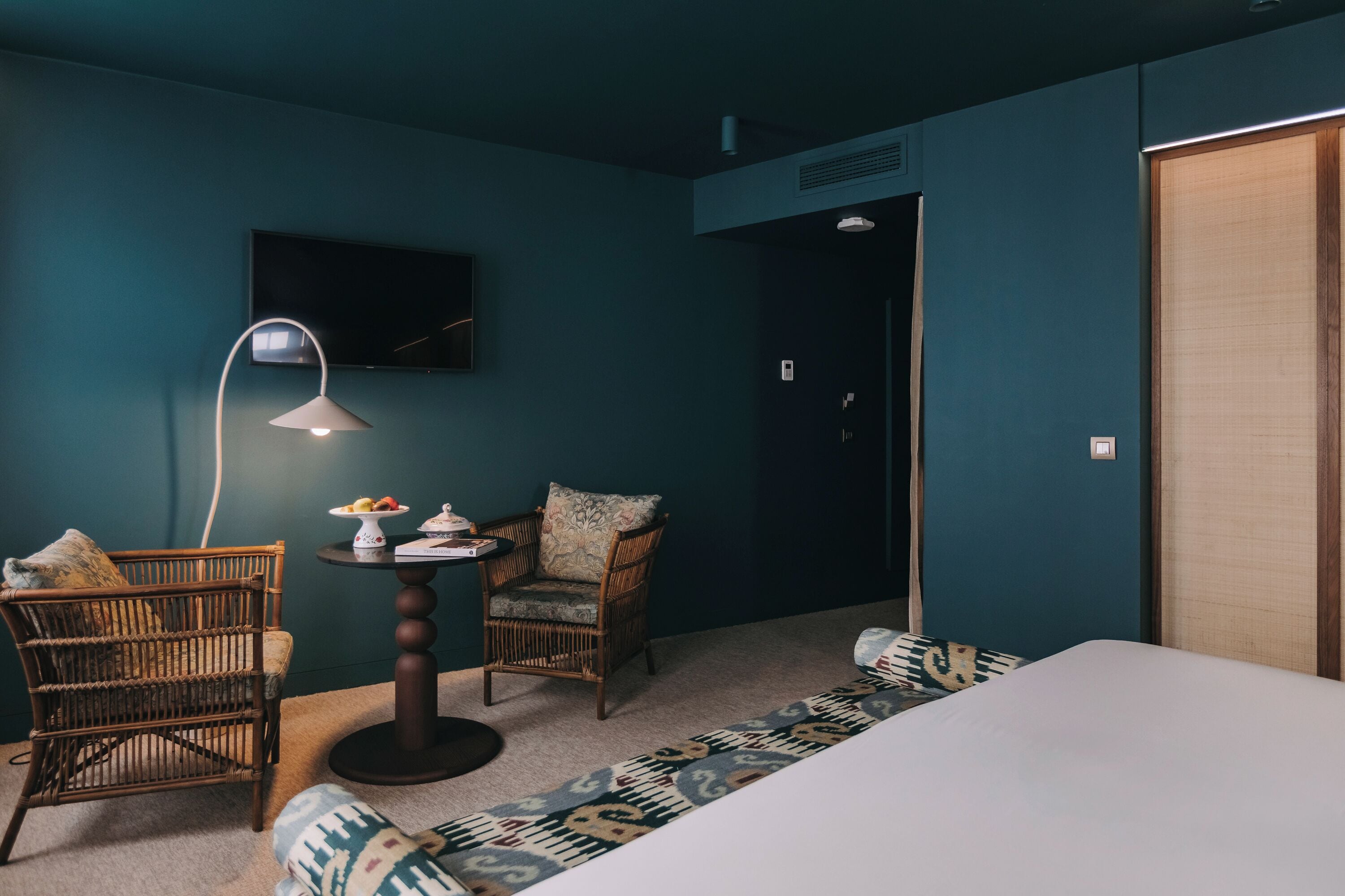 Una habitación con paredes color turquesa, una cama, dos sillones de mimbre, una mesita redonda con una lámpara y un televisor montado en la pared.