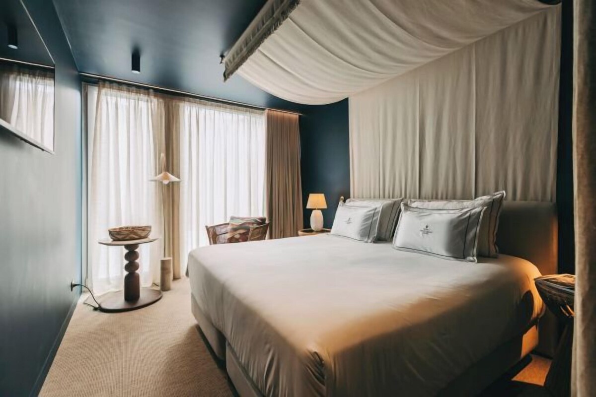 A hotel room with a large bed, a bedside table, a chair, and a window with curtains.