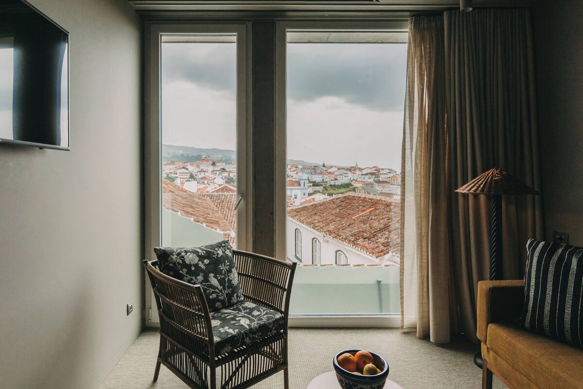 A room with a view of a cityscape through large windows, a wicker chair with a floral cushion, a brown sofa with striped cushions, and a bowl of fruit on the floor.