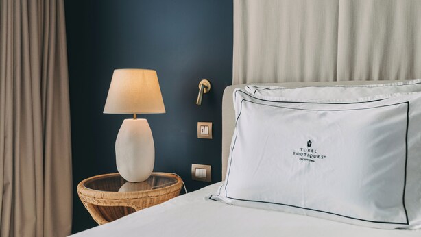 Una habitación de hotel con una cama, una lámpara de noche, una mesita de mimbre e una pared con interruptor de luz y toma de corriente.