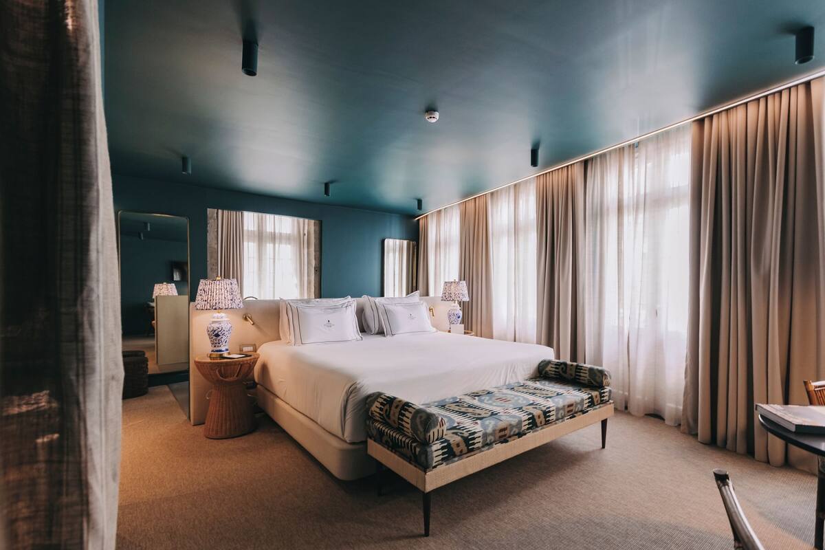 A modern hotel room with a large bed, a bedside table, a lamp, and a view of the outdoors through the window.