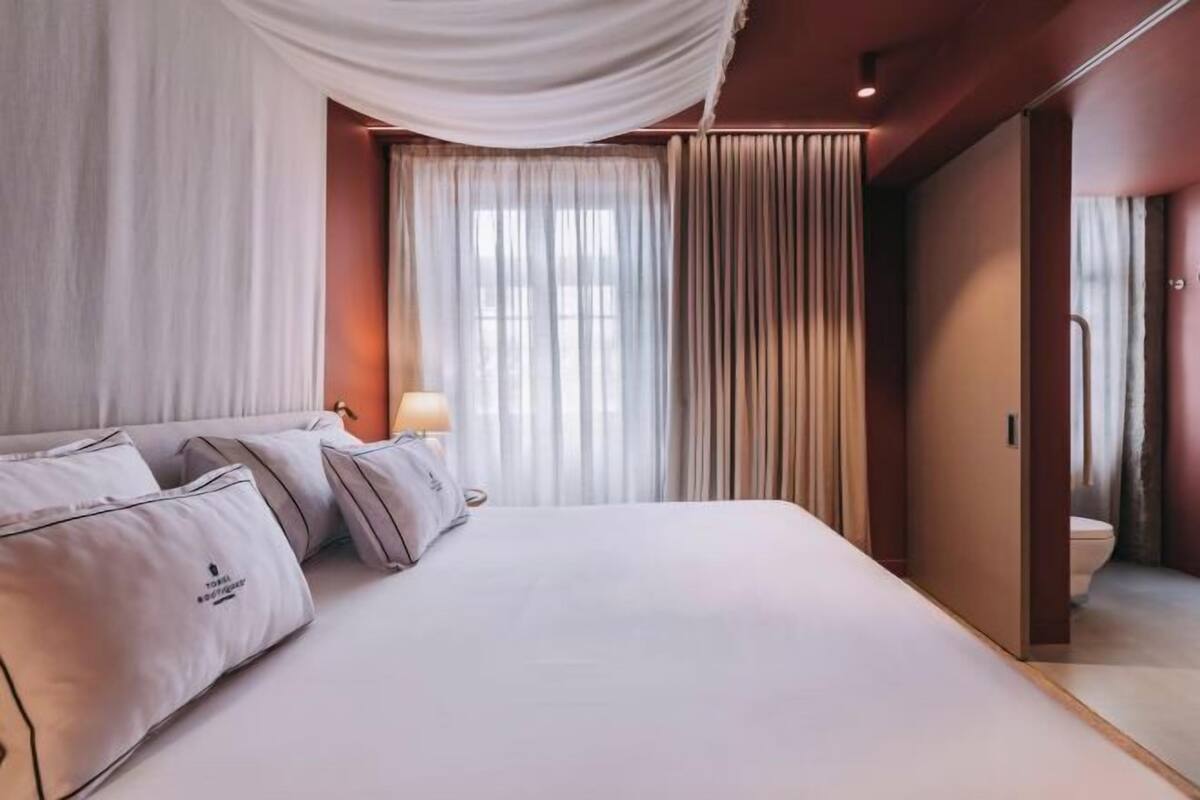 A hotel room with a large bed, a bedside table with a lamp, a window with curtains, and a door leading to a bathroom.