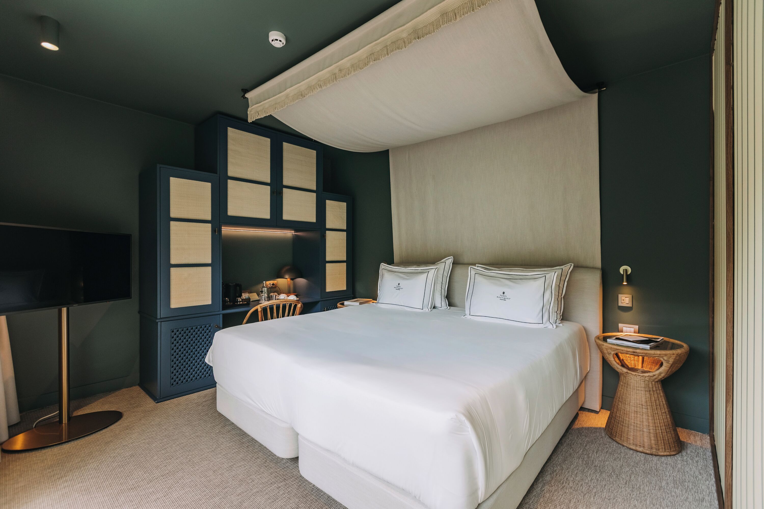 A modern hotel room with a large bed, a television, a desk, and a wicker side table.