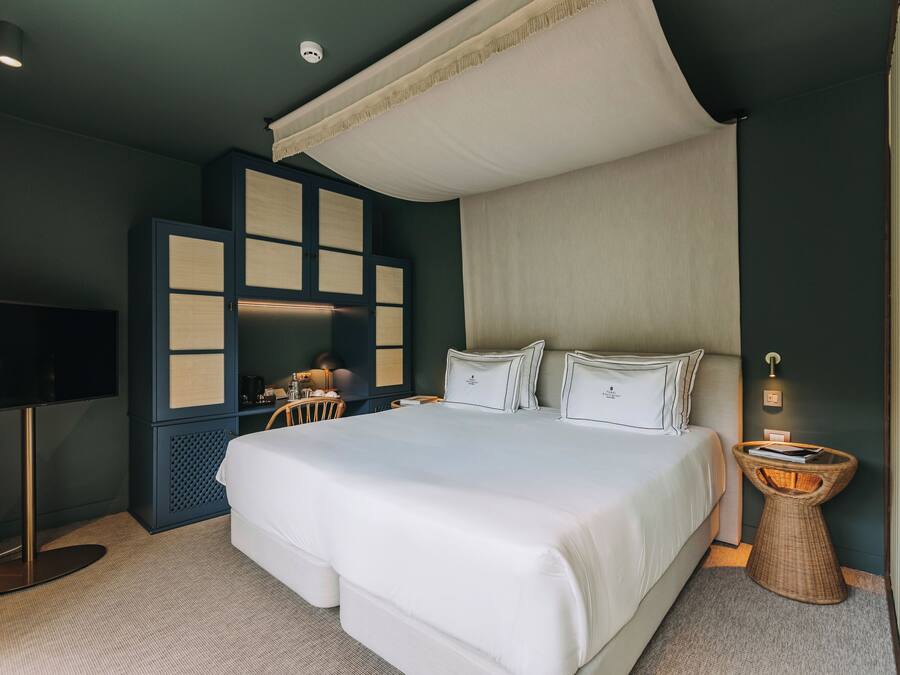 A modern hotel room with a large bed, a television, a desk, and a wicker side table.