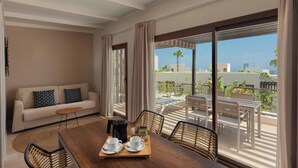 Una habitación de hotel moderna con balcón, una mesa de comedor con cafetera, tazas y bandeja, y un sofá con cojines estampados.