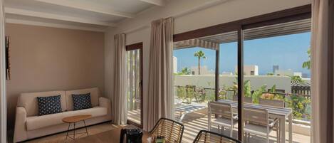 A modern hotel room with a balcony, a dining table with a coffee pot, cups, and a tray, and a sofa with patterned cushions.