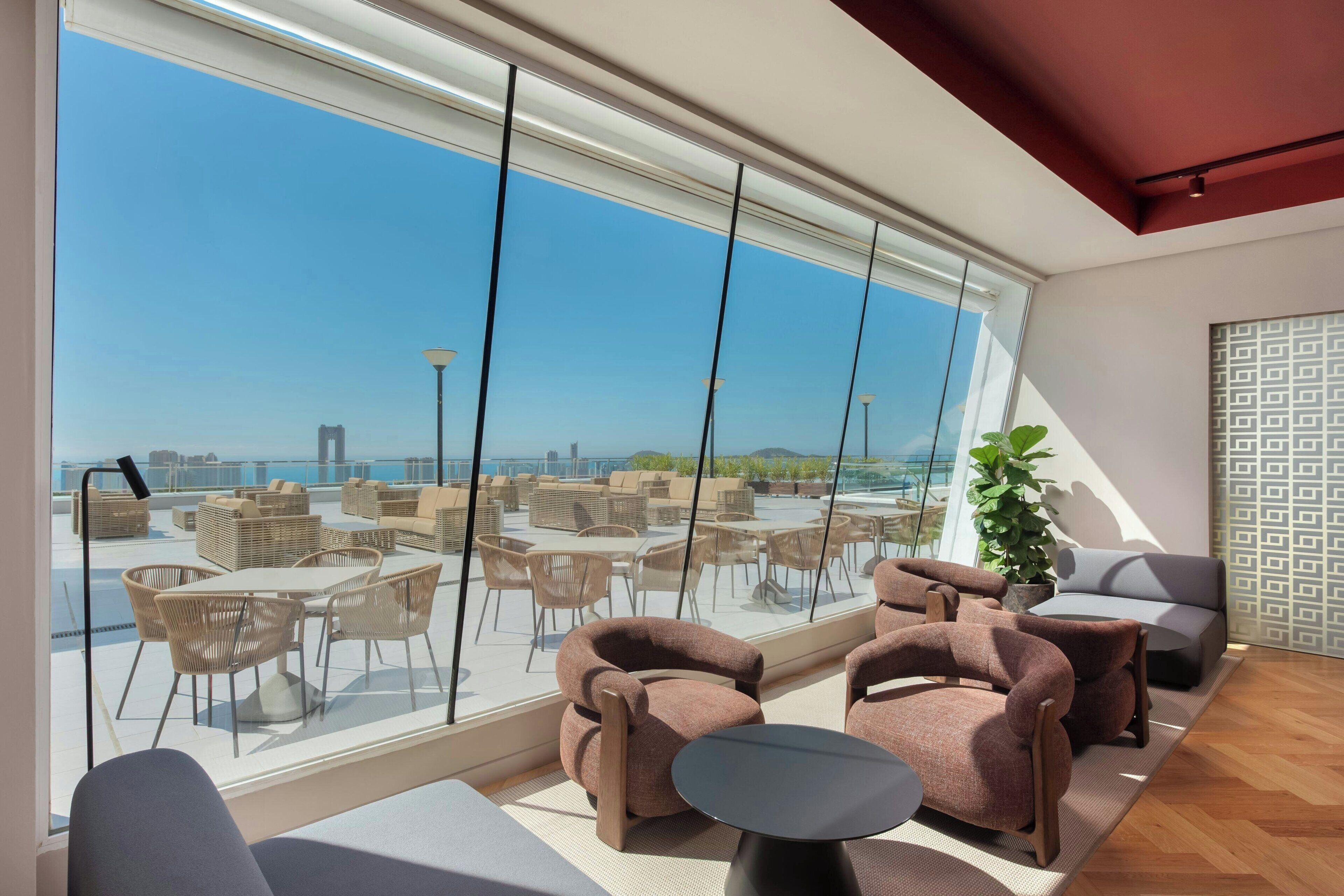A modern interior with large windows overlooking a rooftop bar area.