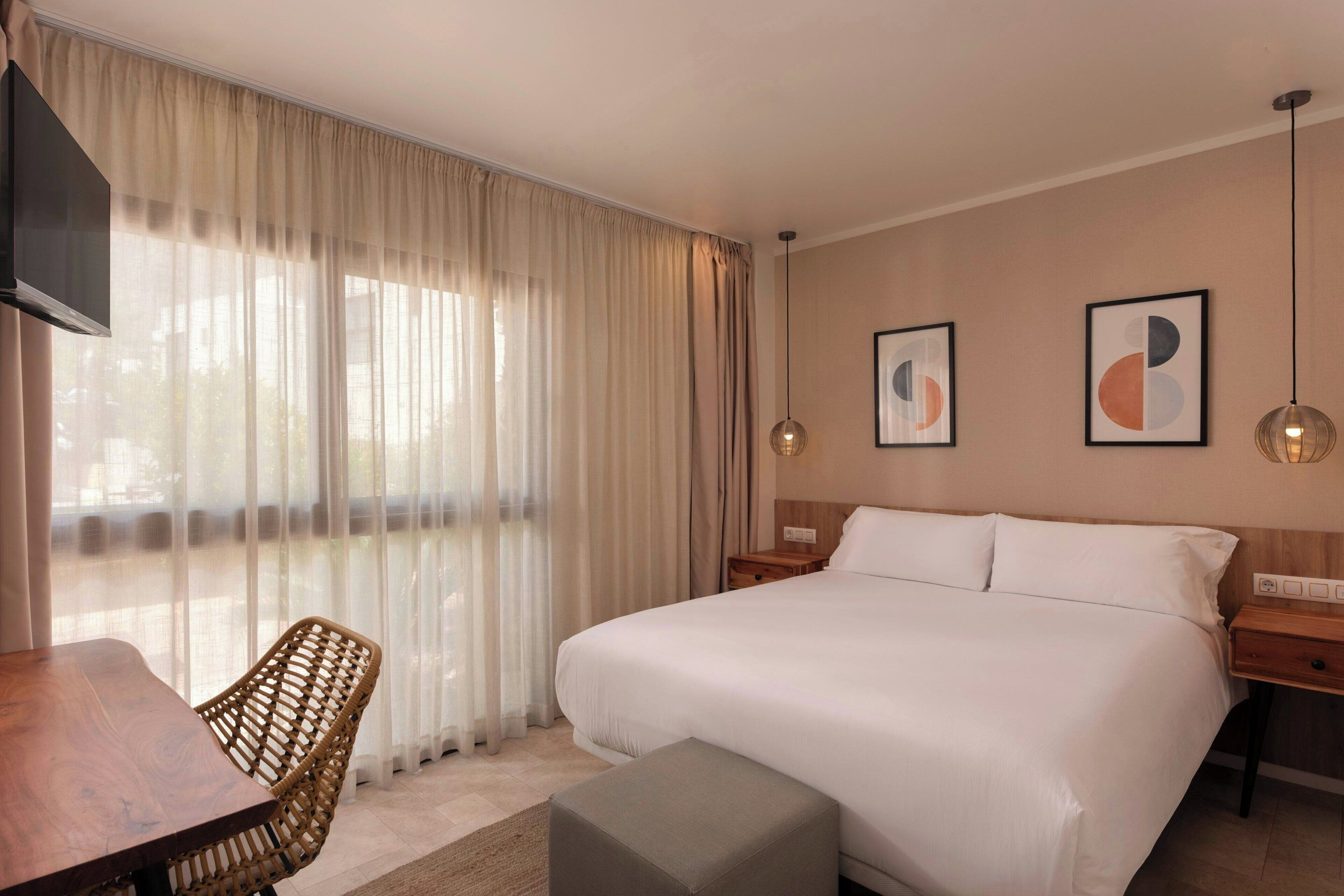 Habitación de hotel con una cama grande, una mesita de noche de madera, una silla de mimbre, una pequeña mesa y un ventanal con cortinas traslúcidas.