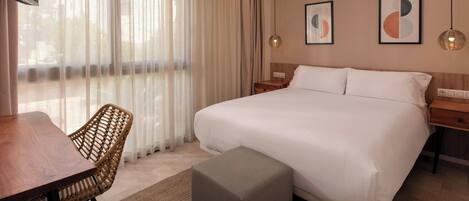 Habitación de hotel con una cama grande, una mesita de noche de madera, una silla de mimbre, una pequeña mesa y un ventanal con cortinas traslúcidas.