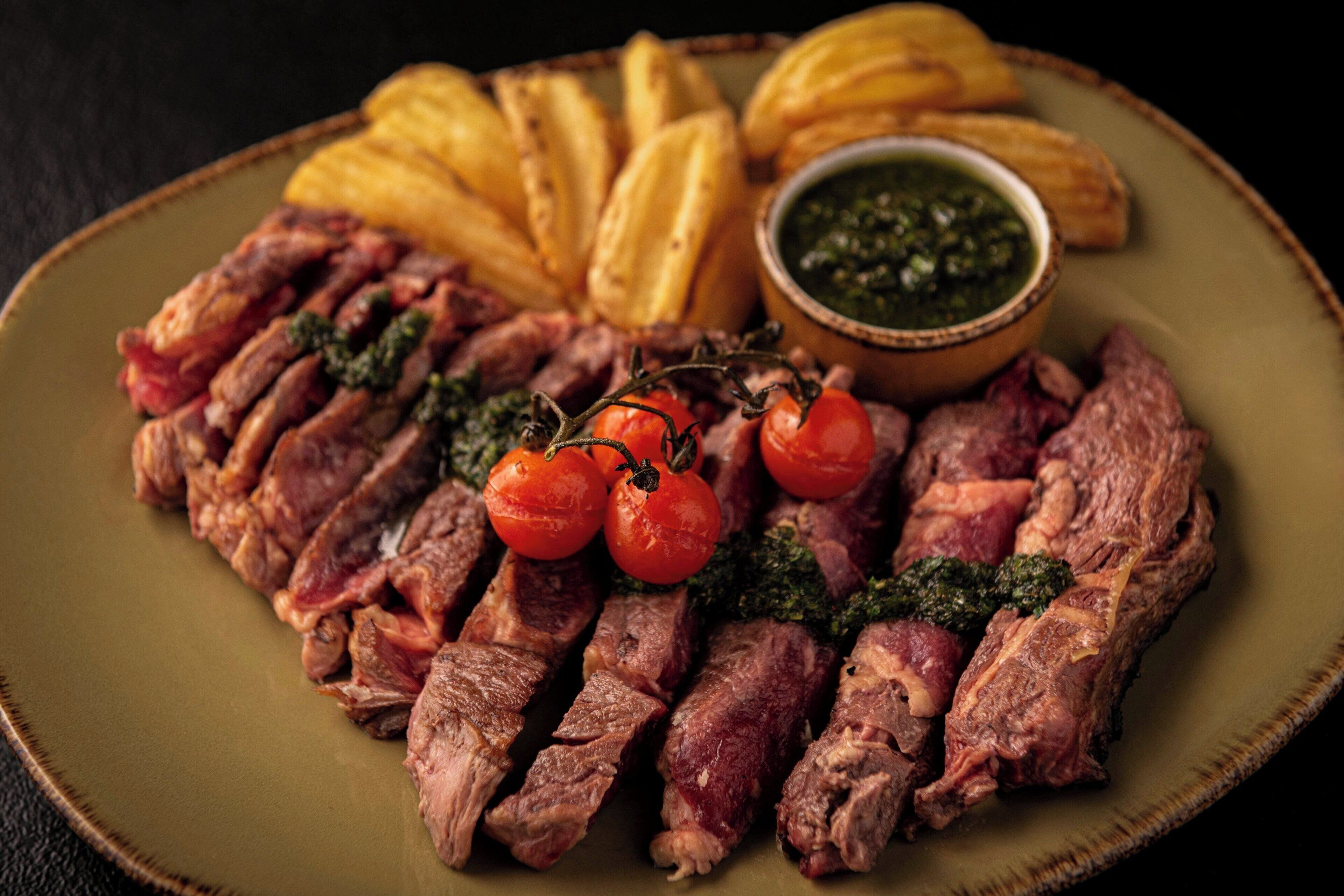 A plate of sliced steak with cherry tomatoes and a bowl of green sauce.