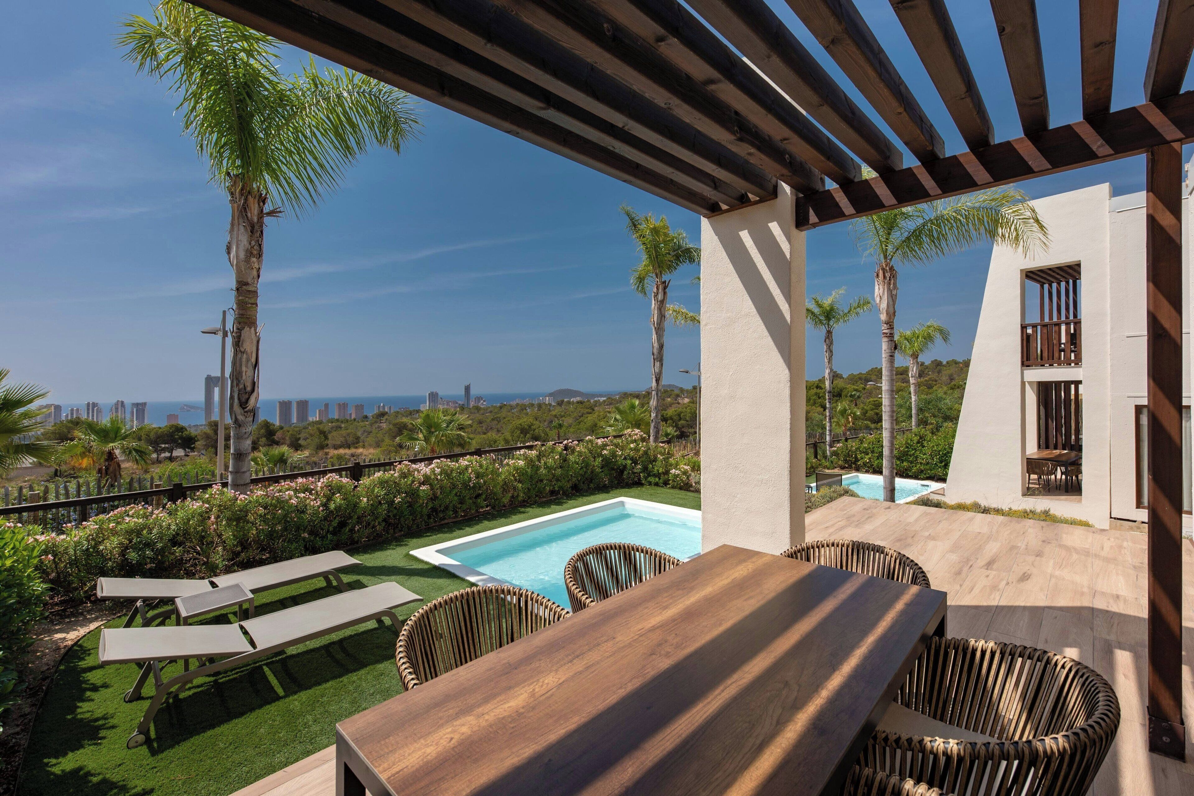 A rooftop pool area with a wooden table and wicker chairs, surrounded by lush greenery and palm trees.