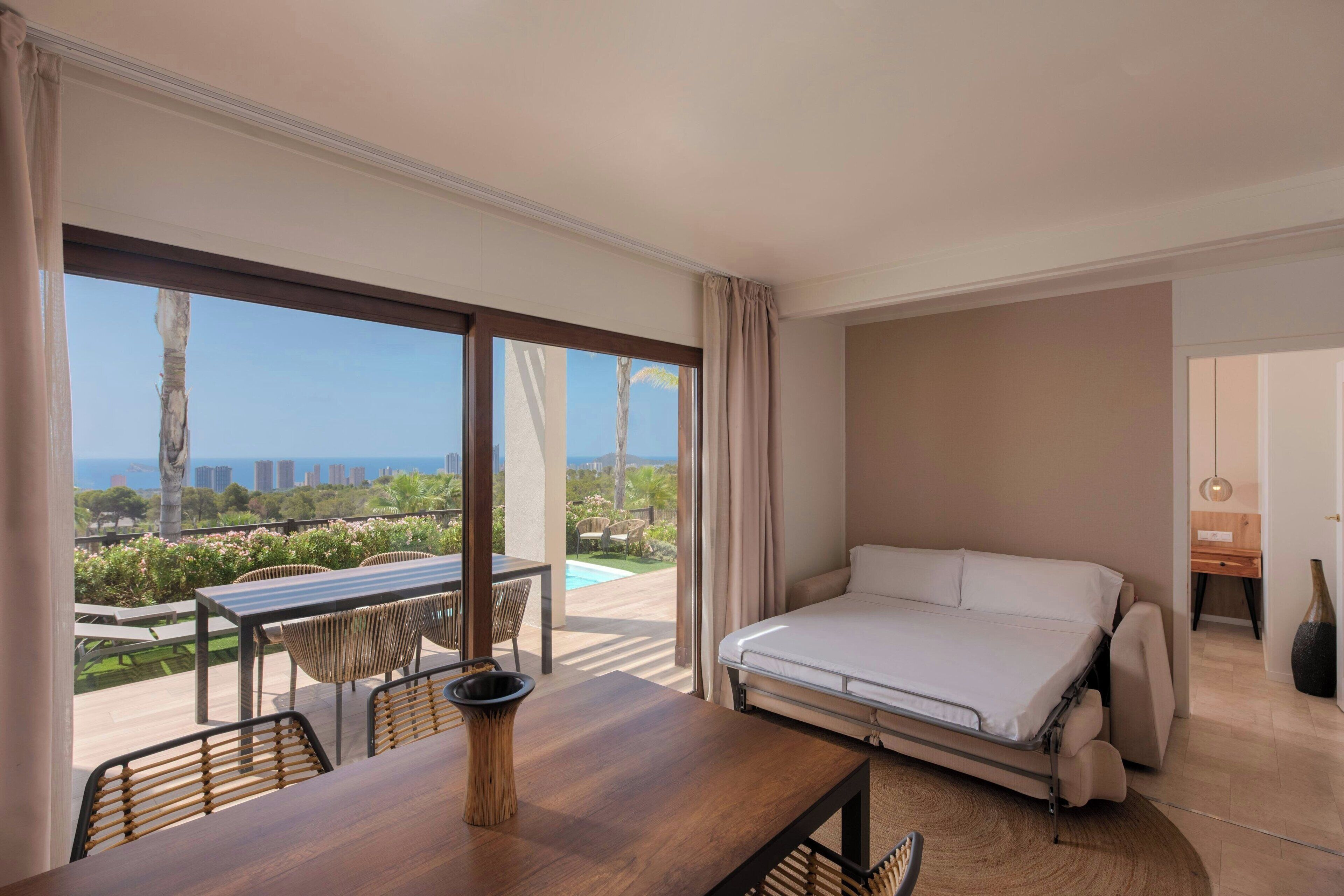 Habitación de hotel con cama, mesa de comedor con sillas y vistas a la ciudad y al océano.
