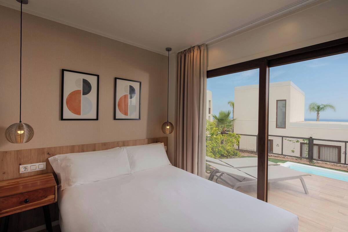 Habitación de hotel con una cama, dos lámparas de pared, una mesita de noche y una ventana con vista a una piscina y edificios.