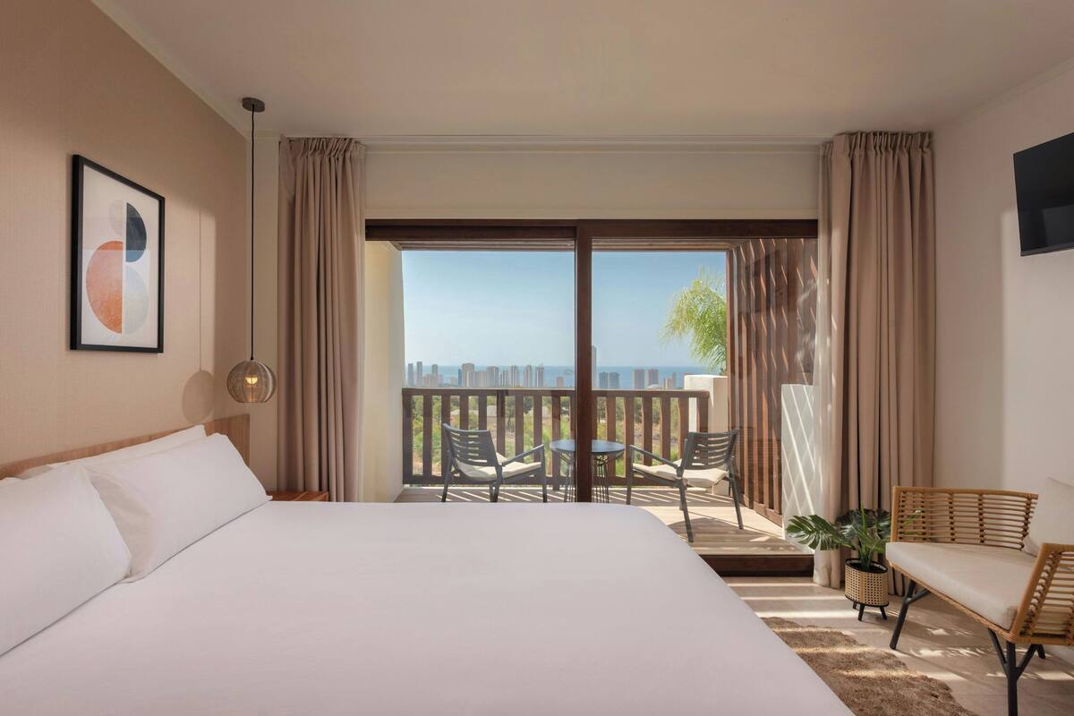Una habitación de hotel con una cama grande, un balcón con mesa y sillas, y un televisor montado en la pared.