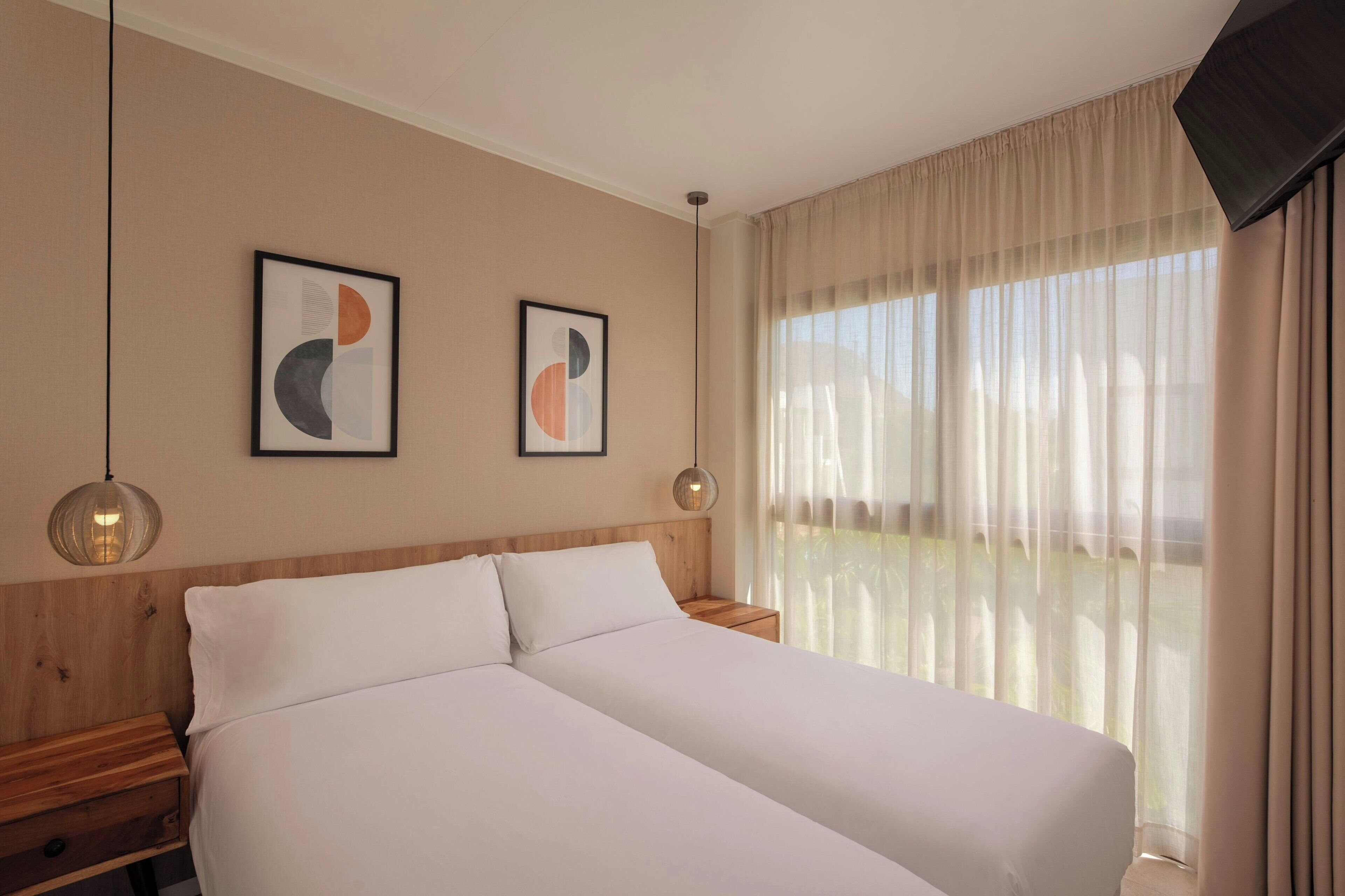 Habitación de hotel con dos camas, cabecera de madera, ventana grande con cortinas translúcidas y dos cuadros abstractos en la pared.
