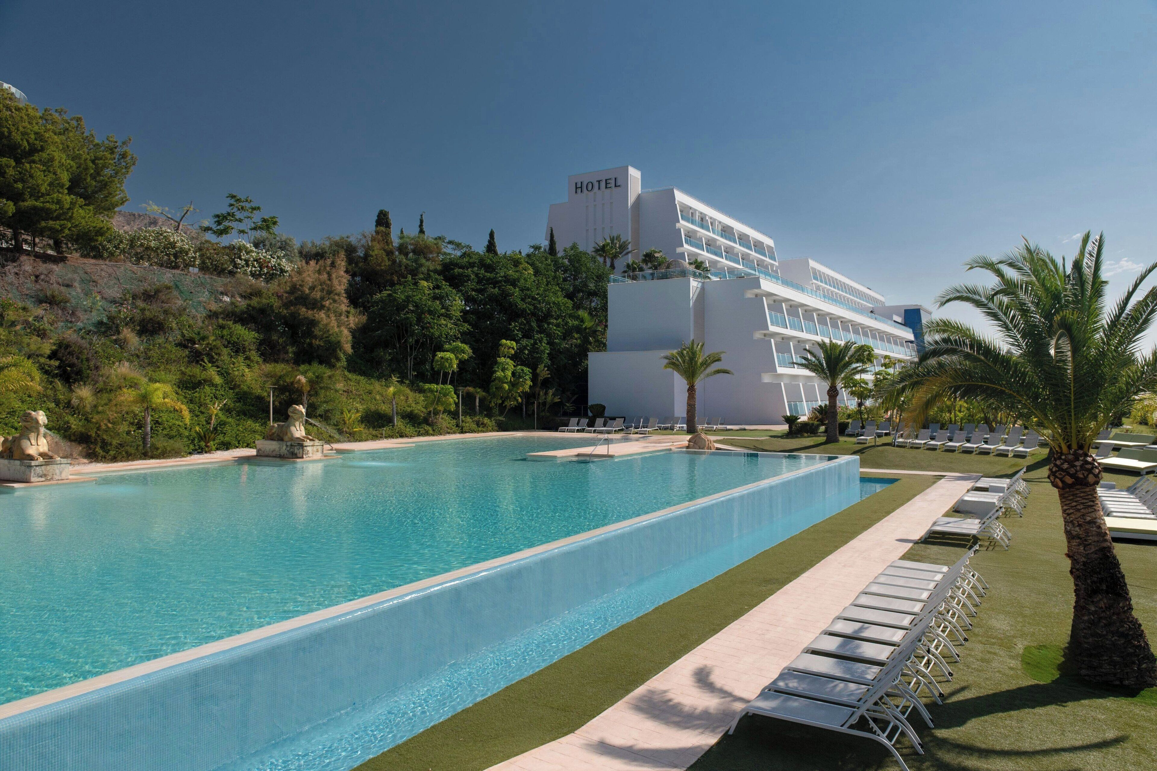 A hotel with a large swimming pool, lounge chairs, and palm trees.
