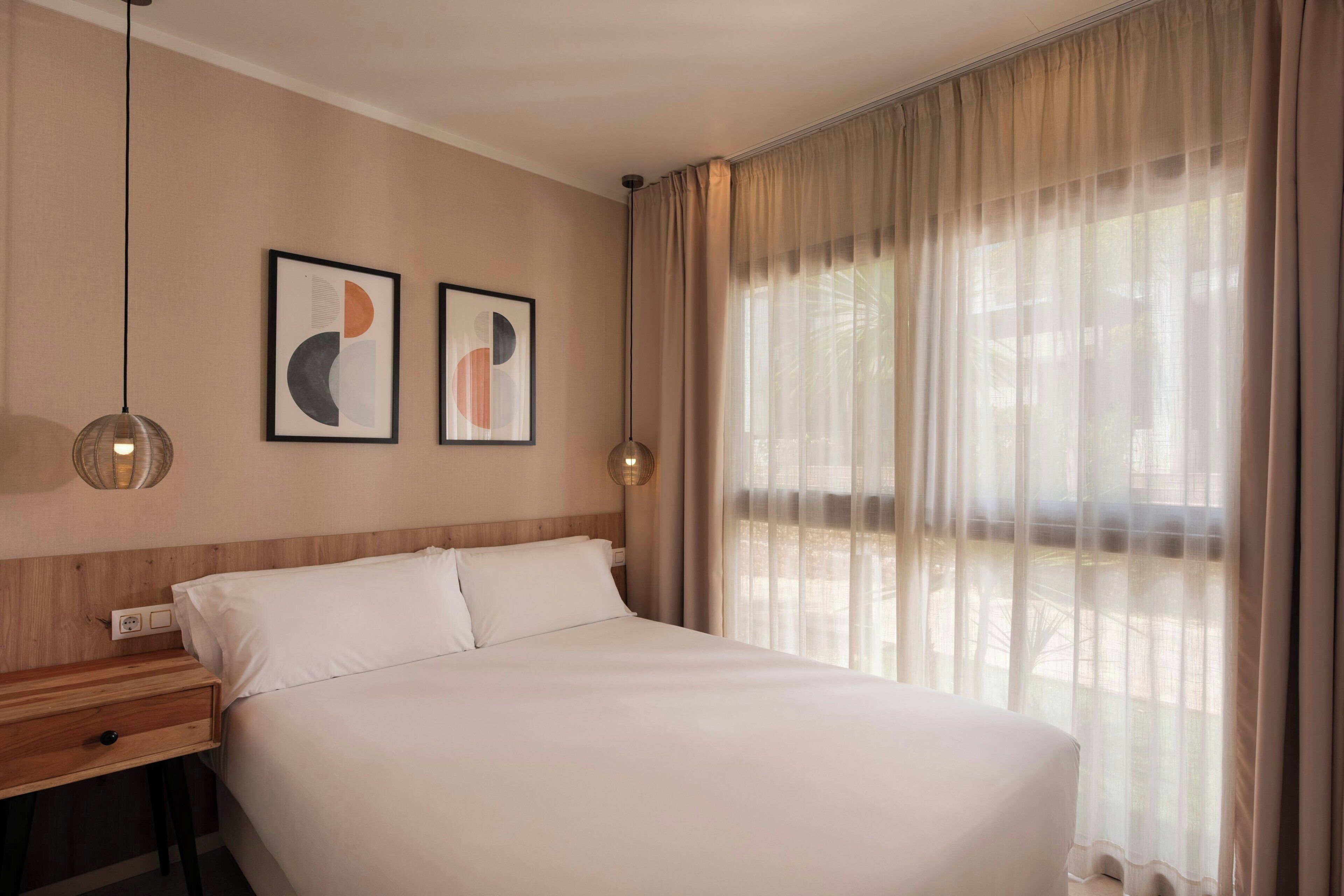 A hotel room with a large bed, two framed abstract artworks on the wall, a bedside table with a lamp, and a window with sheer curtains.