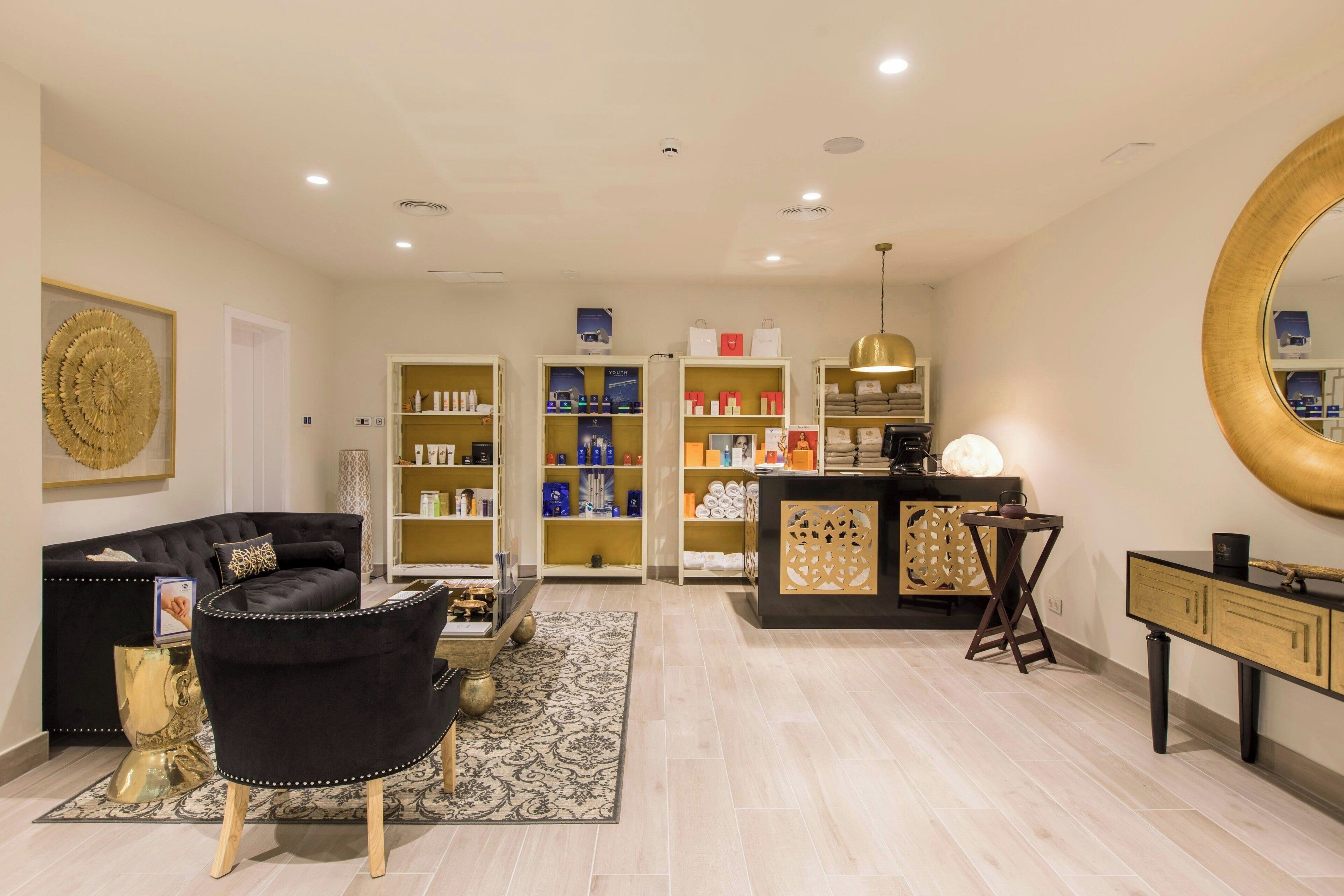 Un salon moderne avec un canapé noir, une table basse et un fauteuil noir. On y trouve des étagères remplies de divers objets et un bureau avec une lampe.