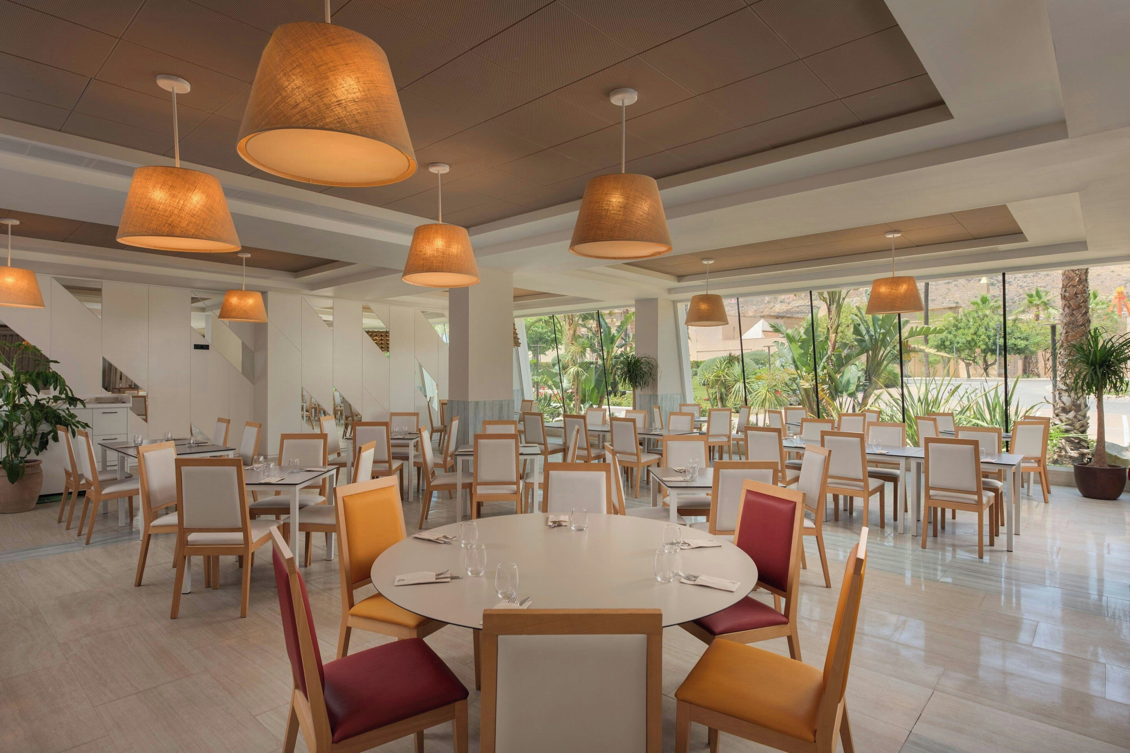 A spacious dining area with round tables and chairs, large hanging lamps, and a view of greenery outside.