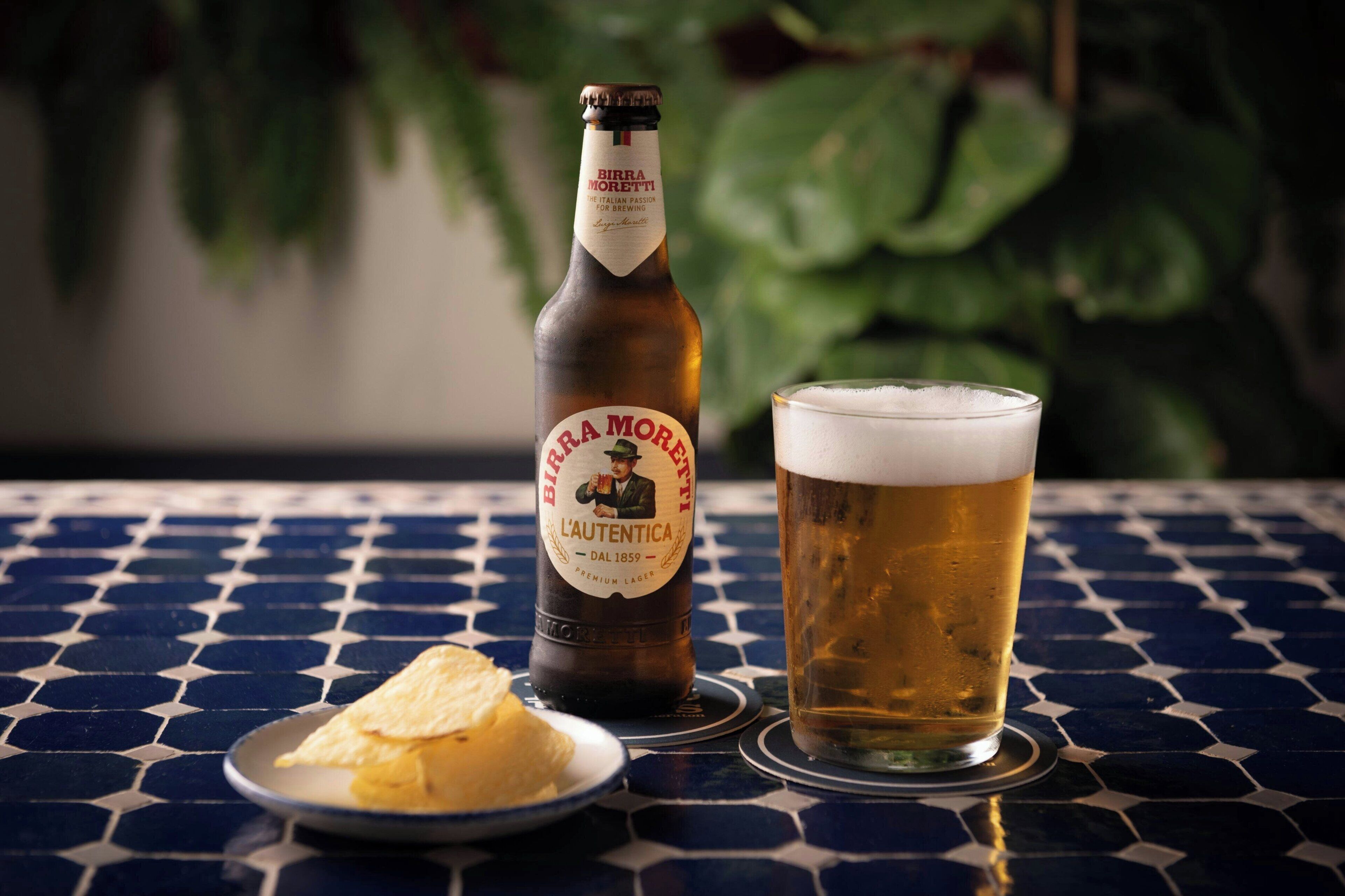 Eine Flasche Birra Moretti L'Authentica Bier, ein Bierglas und ein Teller mit einem Kartoffelchip auf einer gefliesten Tischplatte.