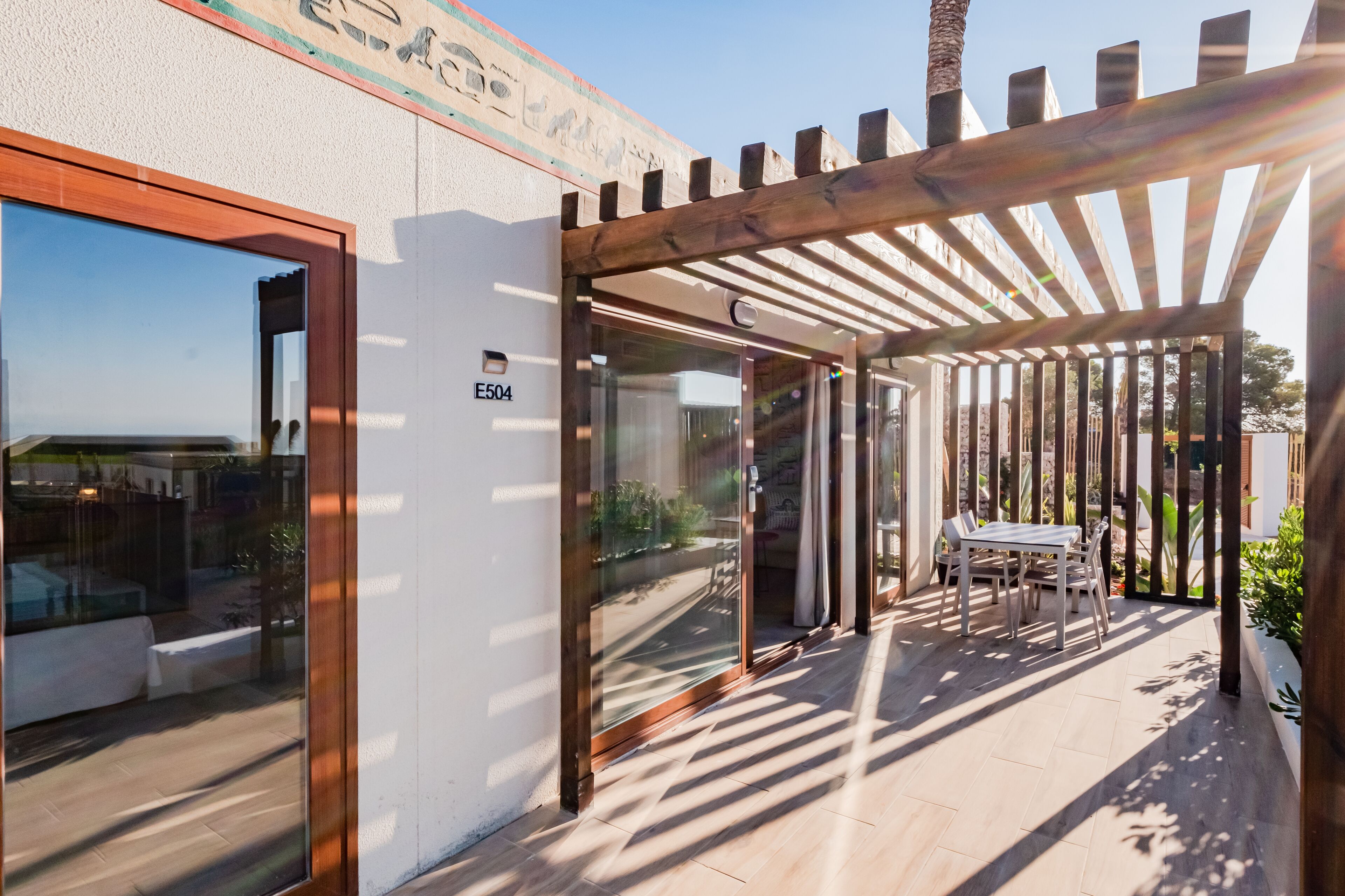 A modern building entrance with a glass door, a wooden pergola, and a patio area with a table and chairs.