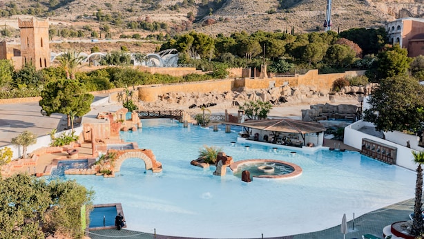 Una gran piscina al aire libre con una fuente, rodeada de sillones de descanso y un paisaje montañoso.