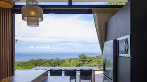 A modern kitchen with a view of the ocean, a dining area with a table and chairs, and a large hanging light fixture. - Luxury modern Villa North Santa Teresa Walk To Surf (Santa Teresa Beach)