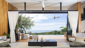 A modern living room with a large sliding glass door leading to a balcony with a view of the ocean. - Luxury modern Villa North Santa Teresa Walk To Surf (Santa Teresa Beach)