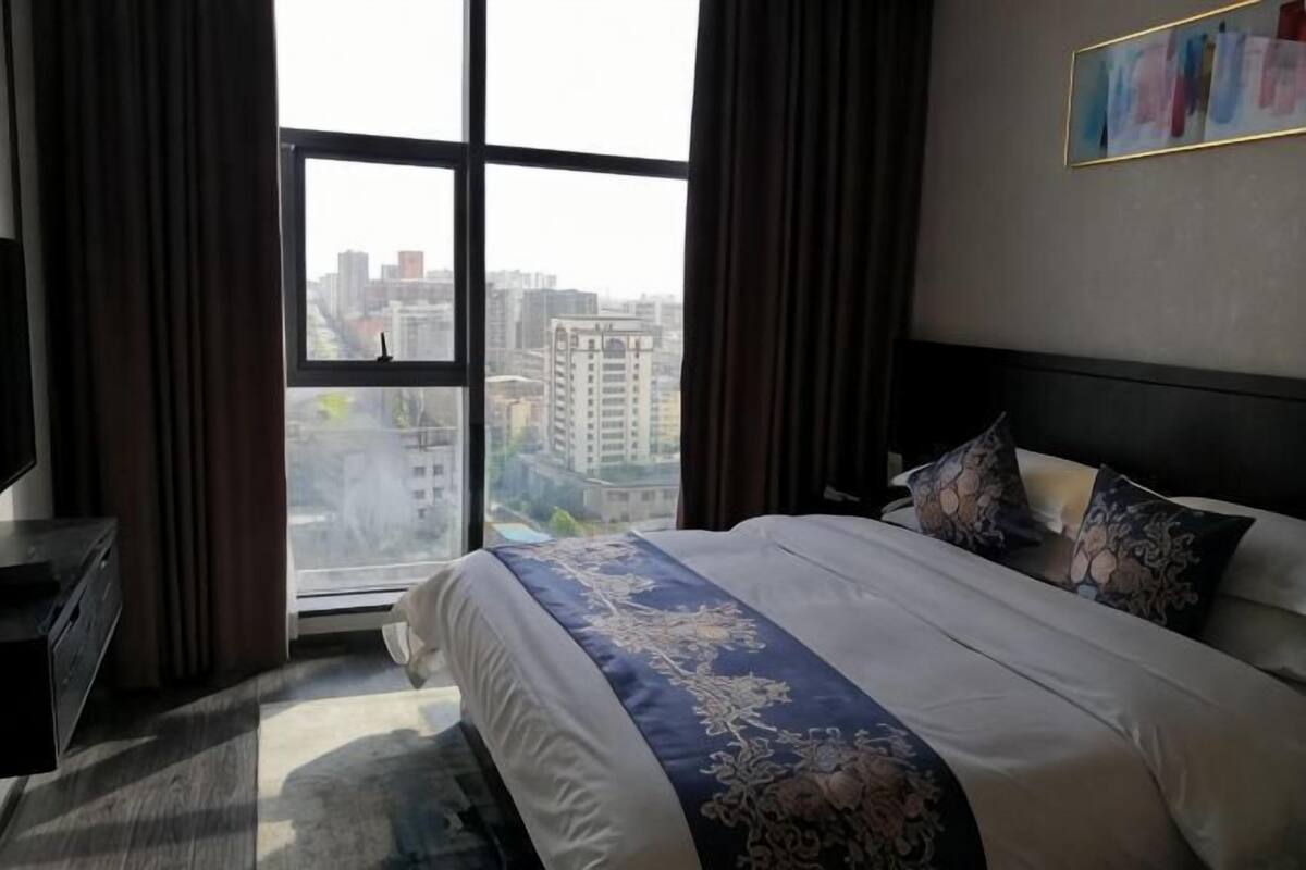 A hotel room with a large bed, a dresser, and a view of the city through a large window.