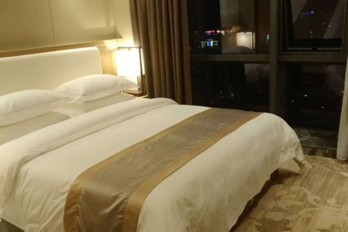 Habitación de hotel con una cama grande, cabecero, mesita de noche y vista nocturna de la ciudad.