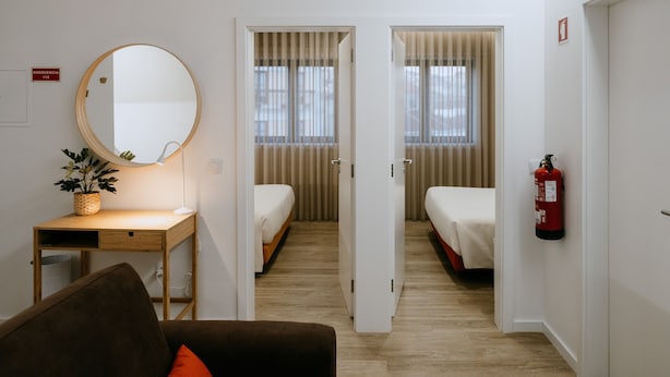 Une chambre d’hôtel avec un bureau en bois, un miroir rond, une petite plante, un lit, une armoire et un extincteur.