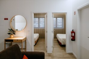 A hotel room with a wooden desk, a round mirror, a small plant, a bed, a wardrobe, and a fire extinguisher.
