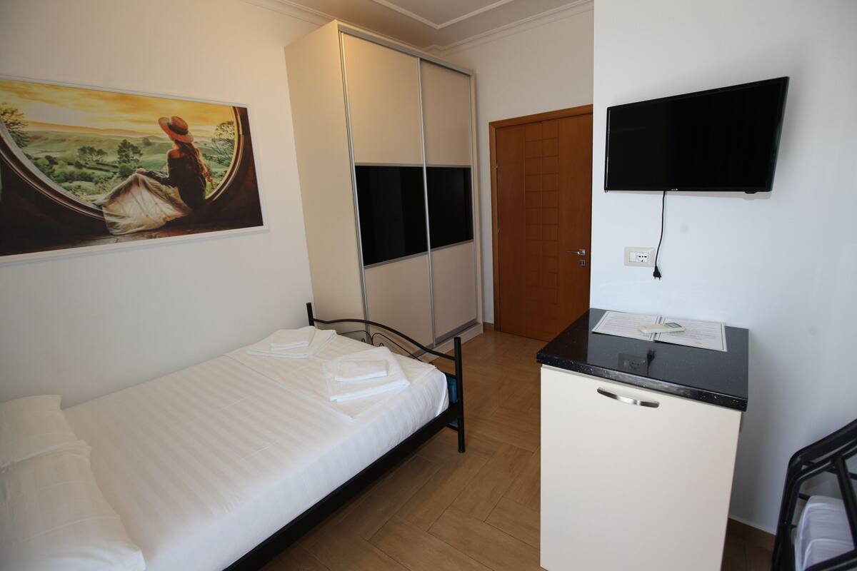 A bedroom with a bed, a wardrobe, a desk, a television, and a painting on the wall.