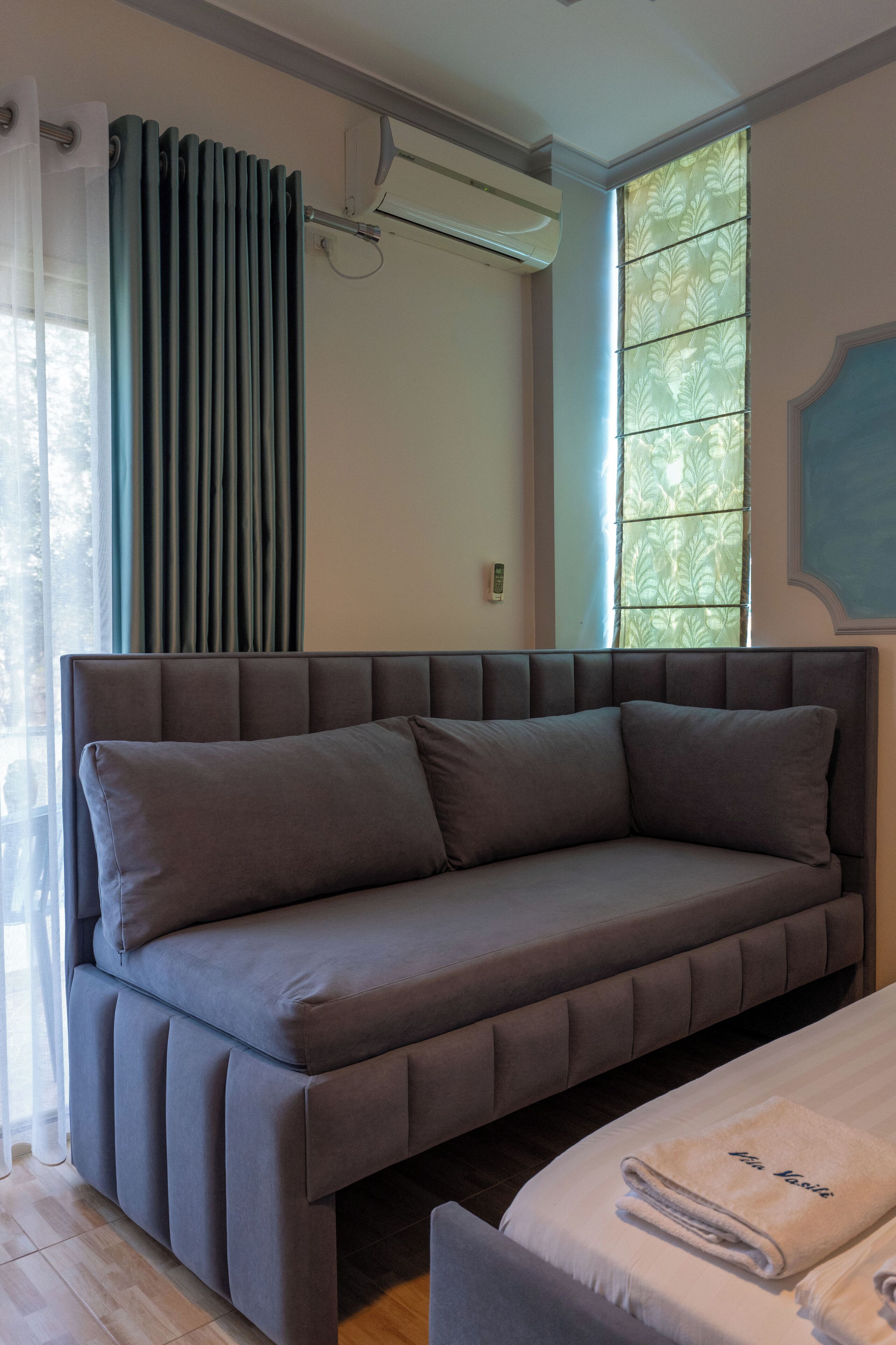 Un dormitorio moderno con un sofá gris, una cama con una toalla doblada, un radiador y una ventana con cortinas estampadas.
