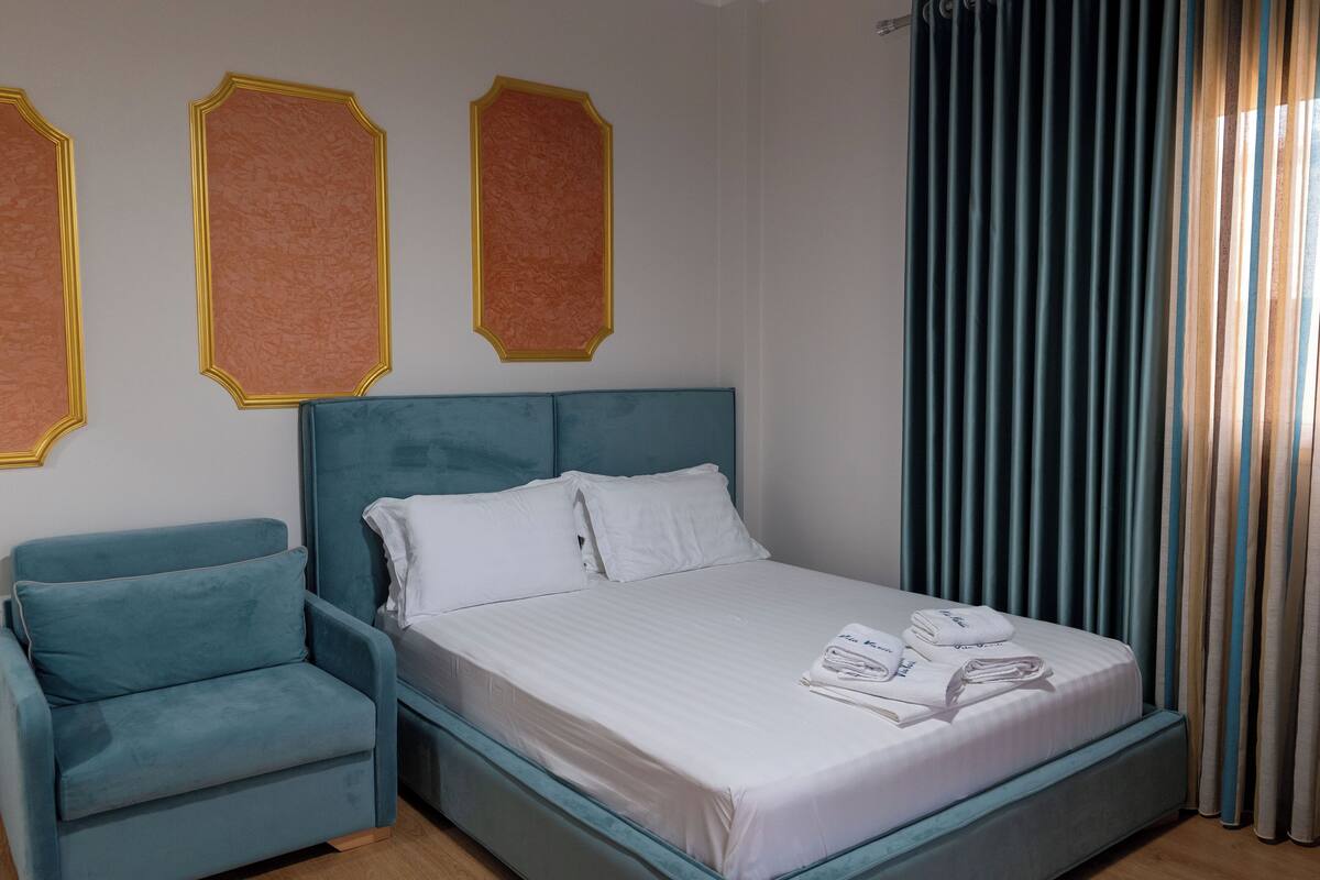 A neatly made bed with white linens and a teal headboard, a teal armchair, and three framed pictures on the wall.