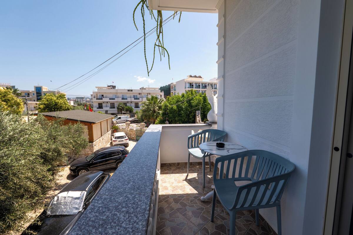 A balcony with a stone floor, two blue chairs, a small table, and a view of buildings and trees.