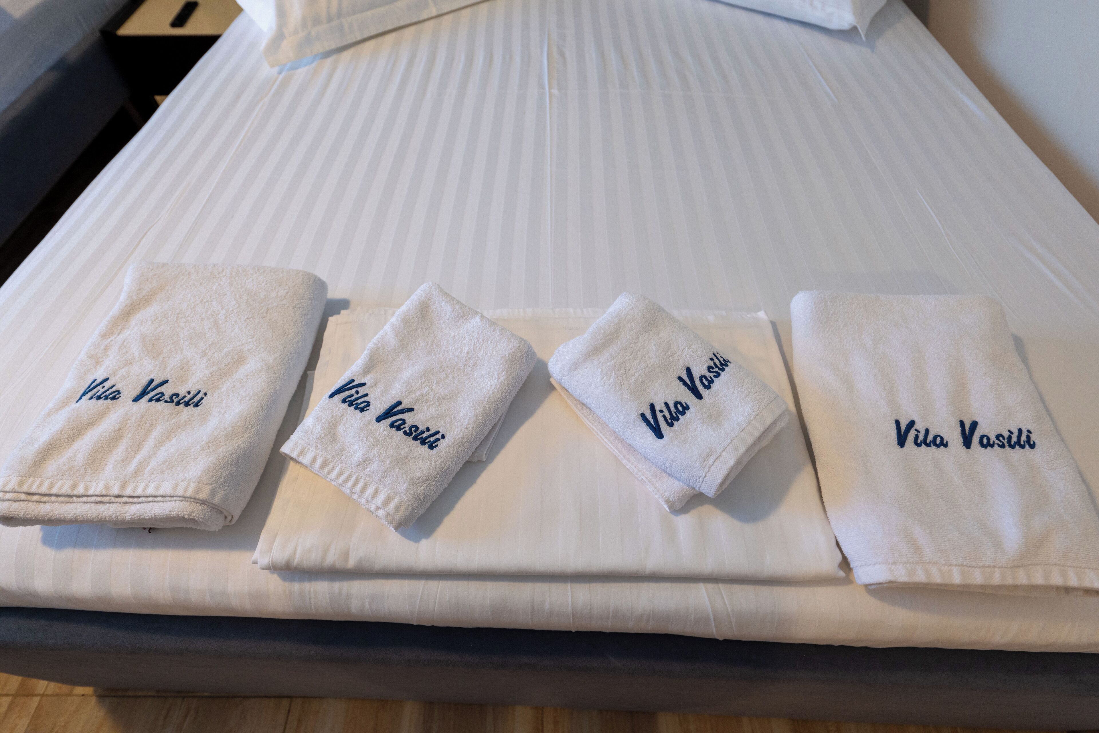 A neatly made bed with four white towels, each with 'Vila Vasili' embroidered in blue.