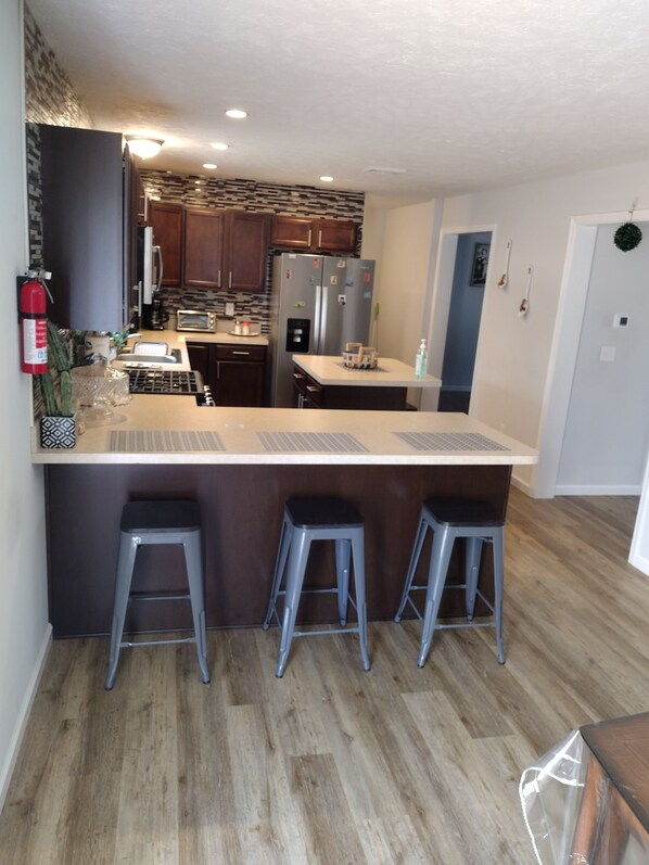 A modern kitchen with a central island, bar stools, wooden cabinets, stainless steel appliances, and a fire extinguisher.