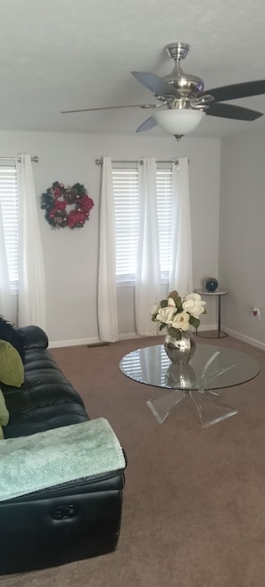 A living room with a ceiling fan, a glass coffee table with a vase of flowers, a black leather couch, and a wreath on the wall.