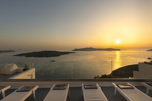 A sunset view from a balcony with lounge chairs and a glass railing overlooking the sea.