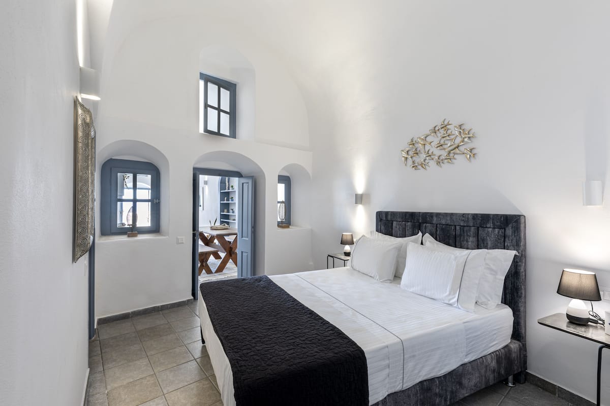 A bedroom with a large bed, a nightstand, a wall-mounted lamp, and a view of the outside.