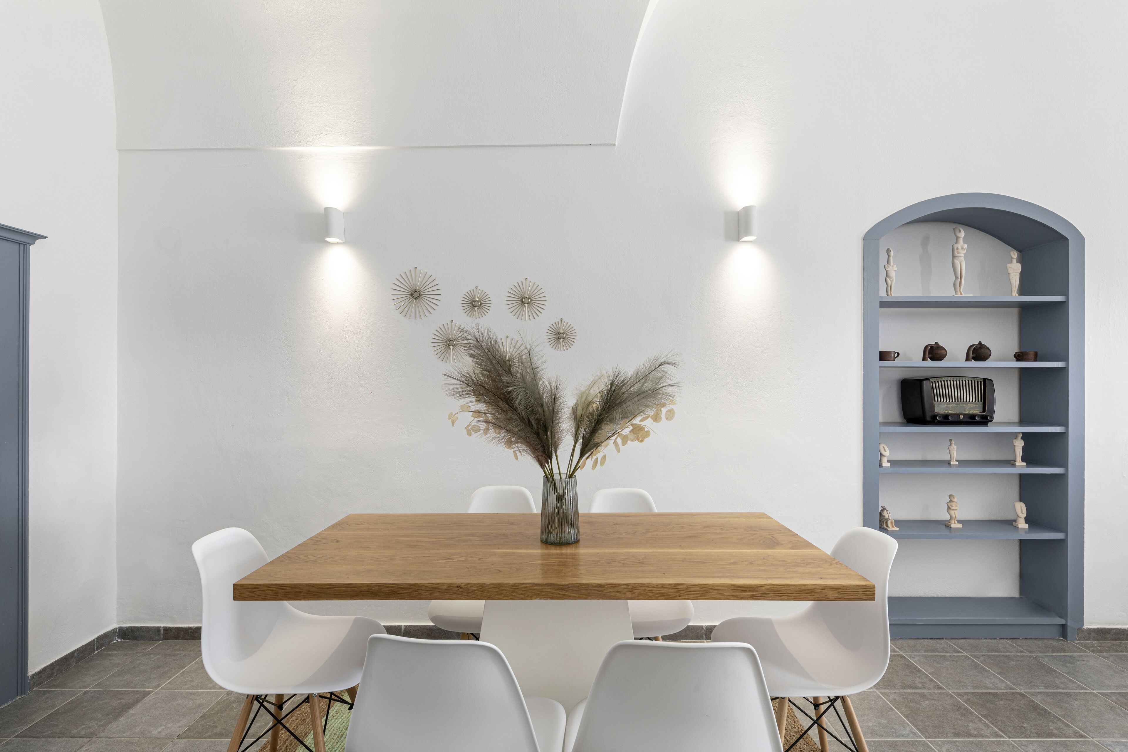 A dining area with a wooden table, white chairs, a vase with dried flowers, wall-mounted lights, and a blue shelving unit with decorative items.